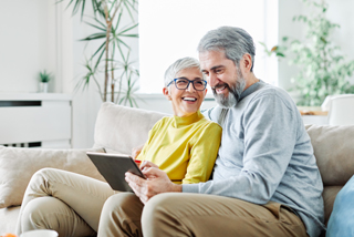 A senior couple sits on a couch in a bright living room, laughing and smiling while looking at a tablet together