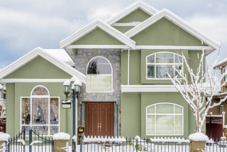 A two-story stucco house painted olive green features a central stone facade and brown double doors