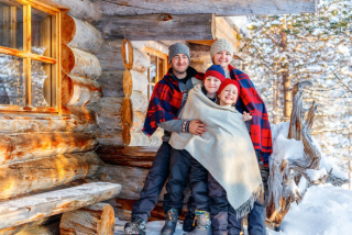 A smiling family of four, wrapped in warm blankets, stands closely together on the snowy porch of a rustic log cabin