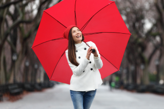 A smiling woman in a white coat and red hat stands on a winter park path, holding a large, bright red umbrella open overhead