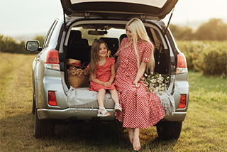 A woman and young girl in matching red dresses sit in the open trunk of a silver car, smiling at each other in a sunny field