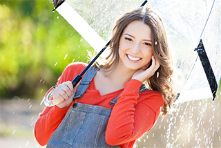 A cheerful young woman in an orange shirt and denim overalls smiles while holding a clear umbrella in the rain